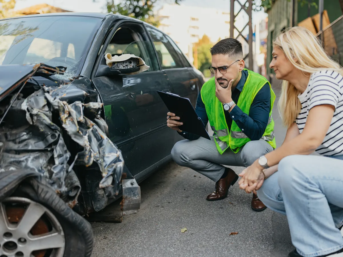 Claims representative with a clipboard talking with a customer beside a car with front-end collision damage